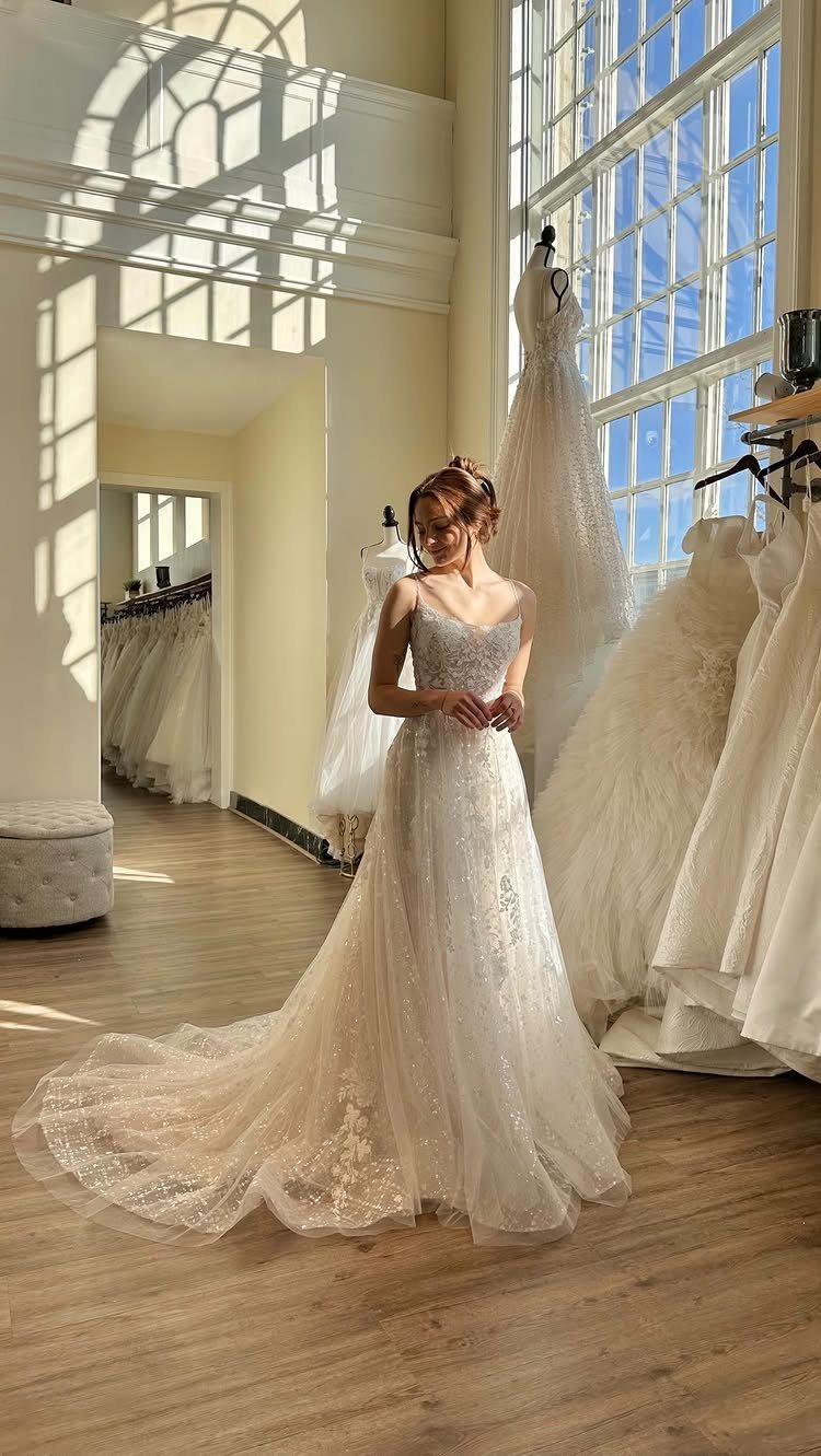 A woman wearing a sparkling white wedding dress with a long train in a sunlit boutique.