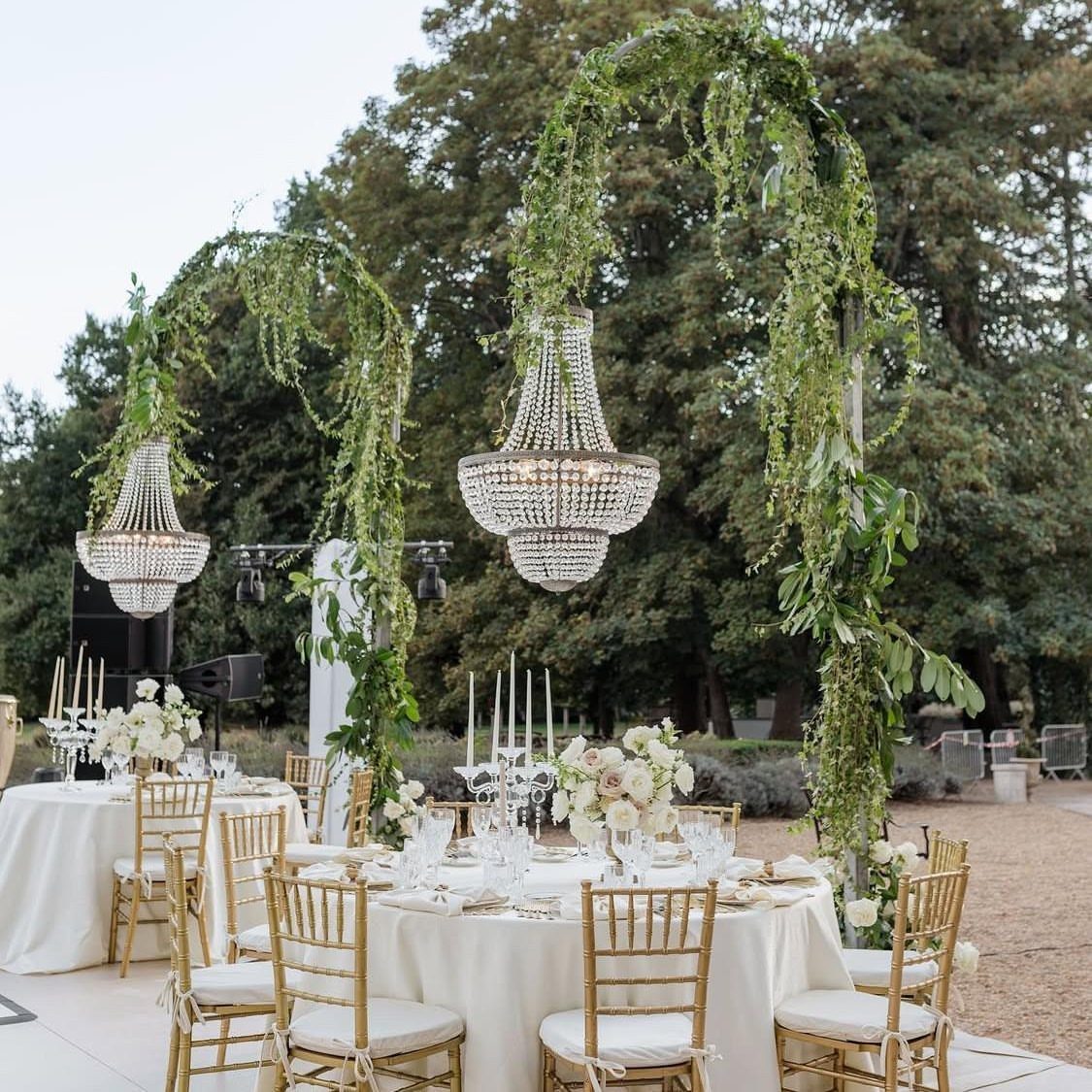 Shabby Chic Wedding: 10 Ideas for Timeworn Charm 3 Crystal chandeliers hanging from lush green floral arches over elegant outdoor wedding reception tables.