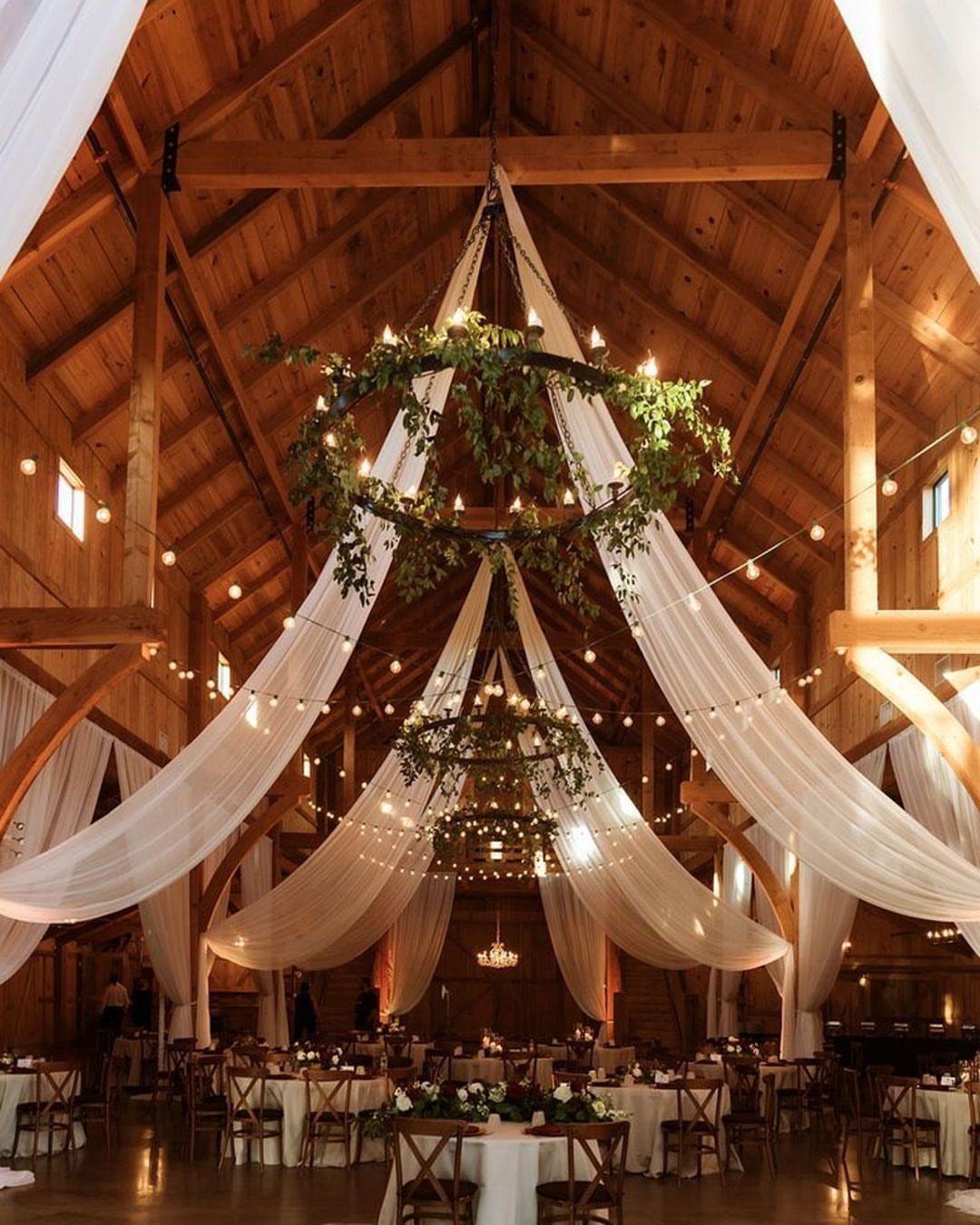 Shabby Chic Wedding: 10 Ideas for Timeworn Charm 6 A decorated wooden barn interior with white fabric draping, greenery chandeliers, and wedding reception tables.