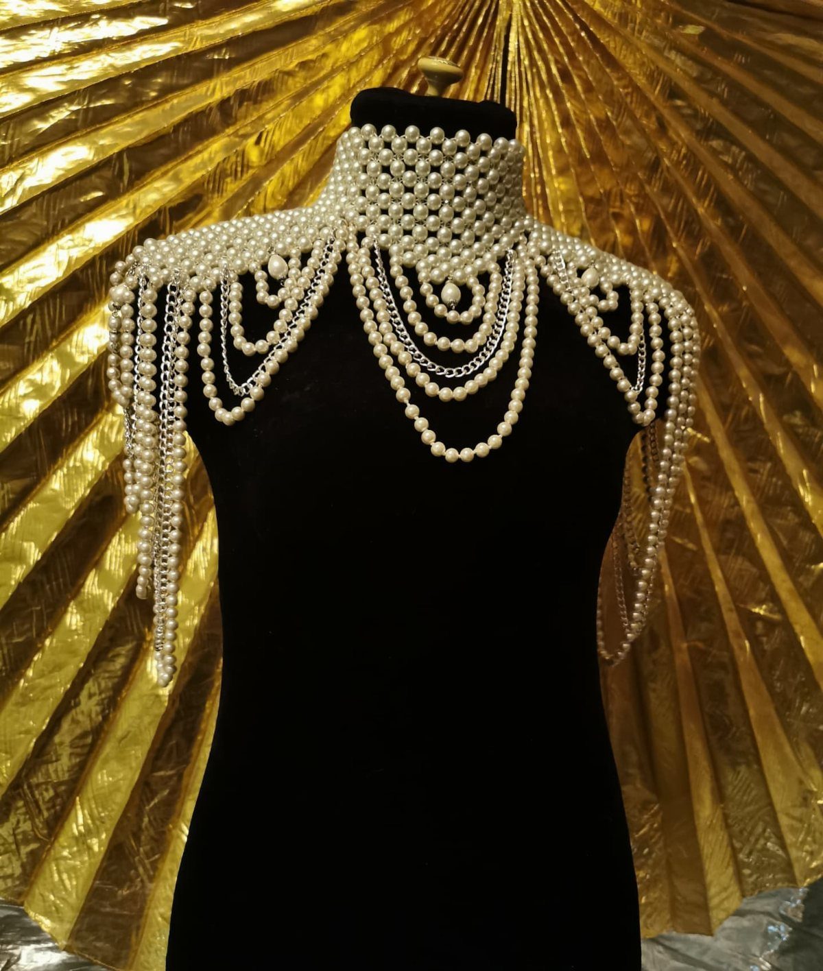 A multi-strand pearl shoulder necklace draped over a black velvet mannequin bust against gold pleats.