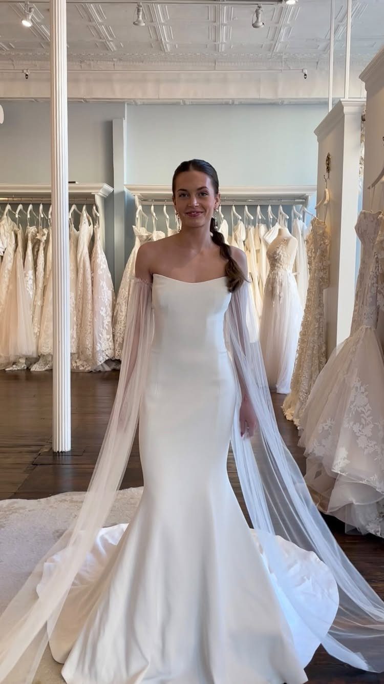 A woman posing in a white strapless mermaid wedding dress with long sheer fabric sleeves.