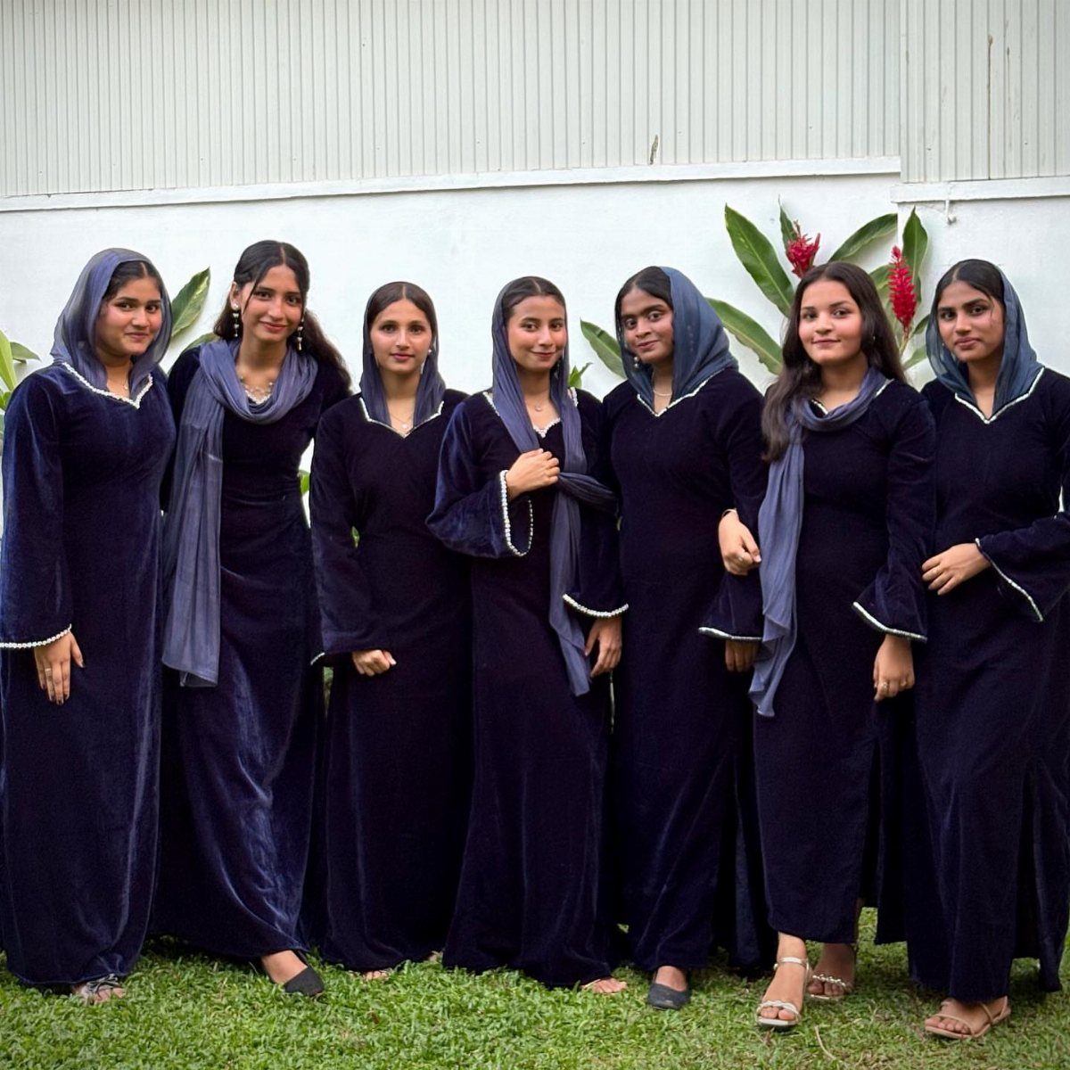 Seven women standing together wearing long navy blue velvet dresses and grey scarves outdoors.