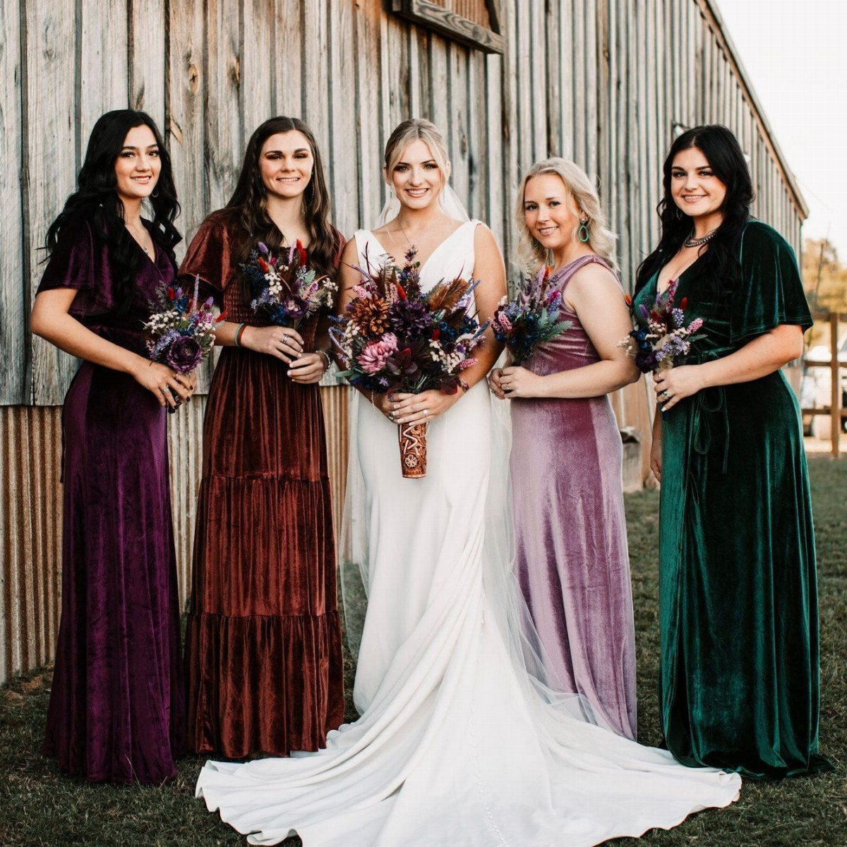 Four bridesmaids in colorful velvet dresses stand with a bride in front of a wooden barn.