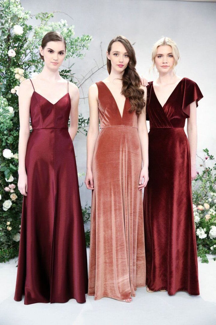 Three women posing in long burgundy, rust, and dark red formal dresses against a floral background.