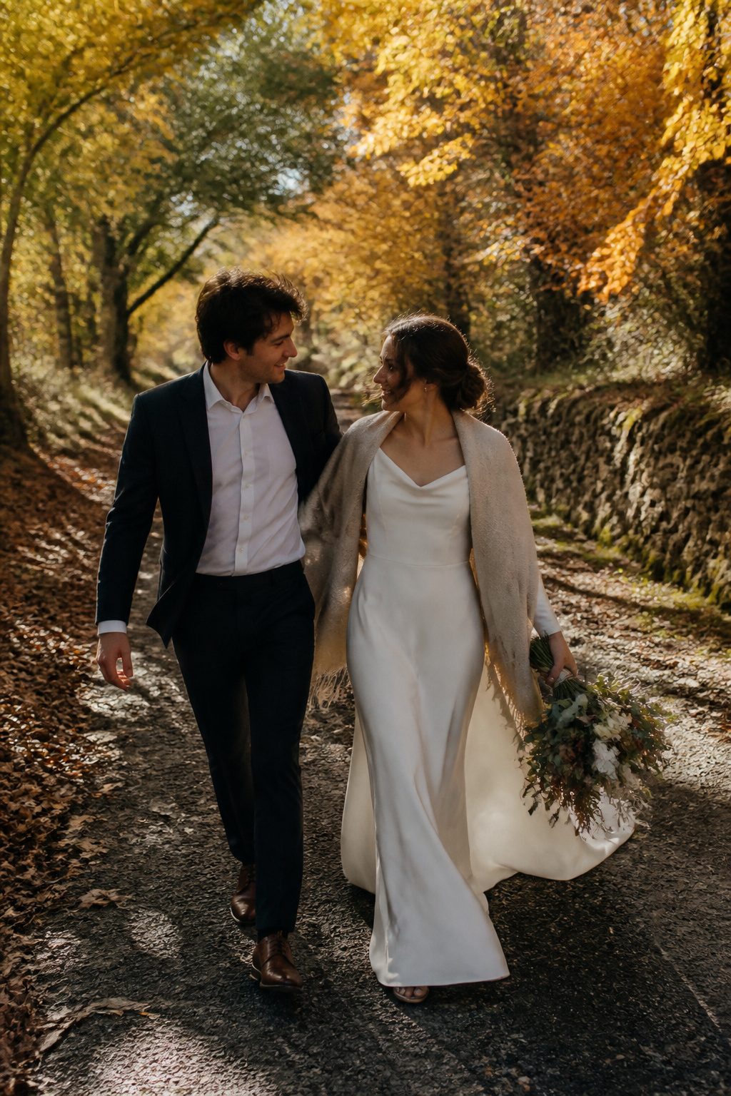Wedding Couple Photos: Ideas, Planning & Timing in 2026 4 A couple walking down a path surrounded by autumn foliage.