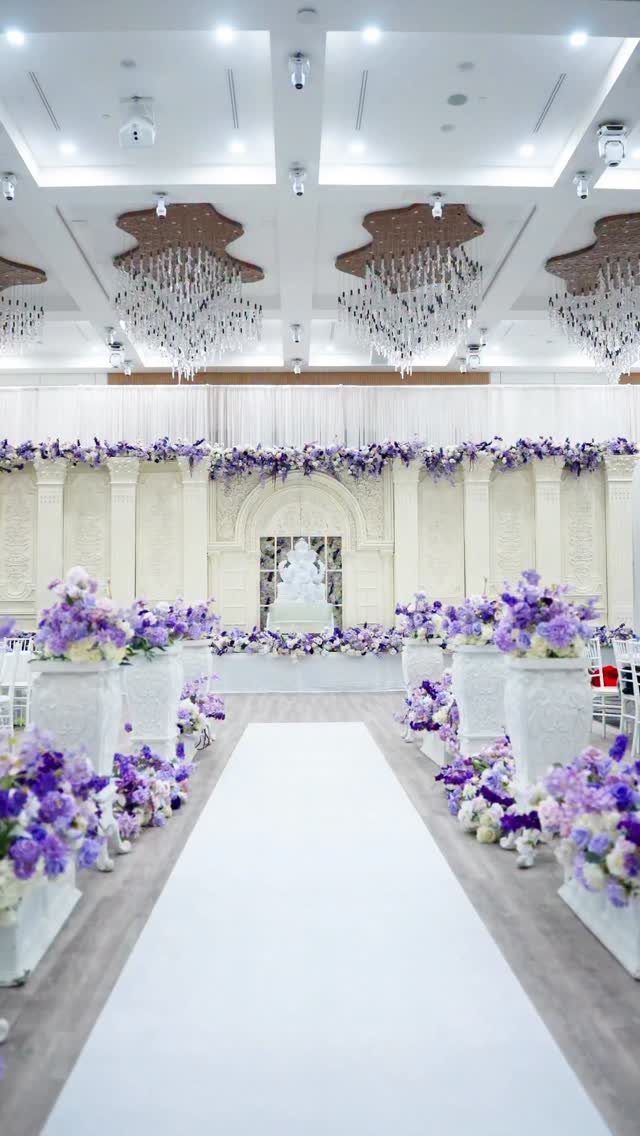 White wedding aisle runner lined with large pedestals of purple and lavender flowers.