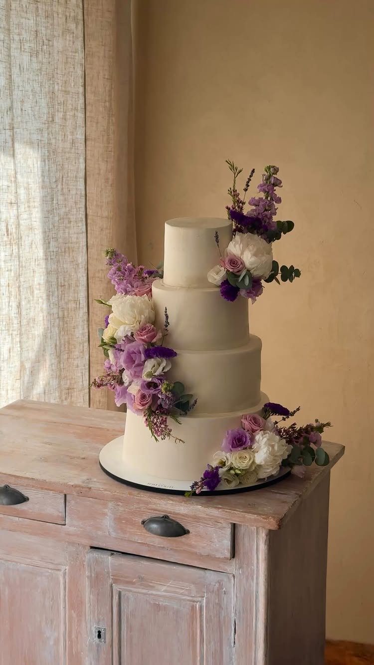 A three-tiered white wedding cake adorned with purple flowers and lavender sprigs on a wooden sideboard.
