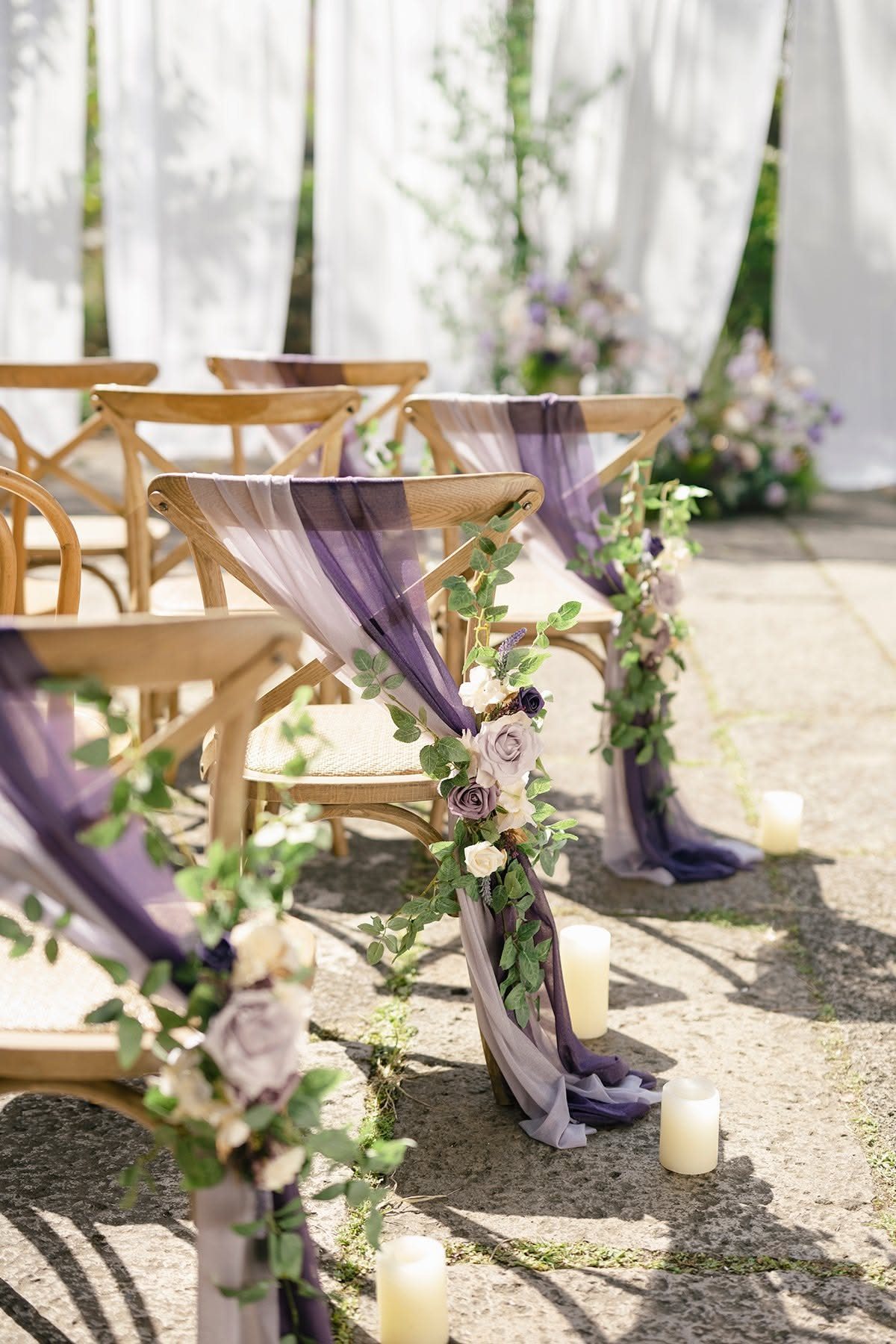 Wooden chairs decorated with purple fabric and floral greenery arranged on a stone patio.