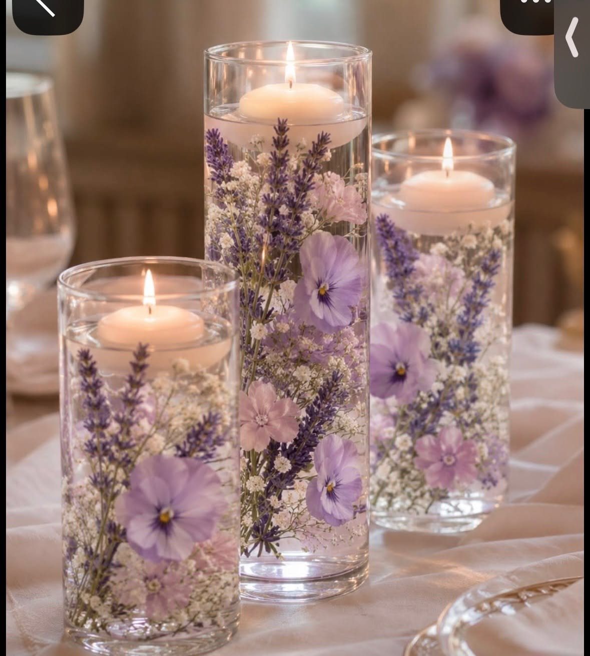 Three glass vases containing lavender, purple flowers, and lit floating candles on a white tablecloth.