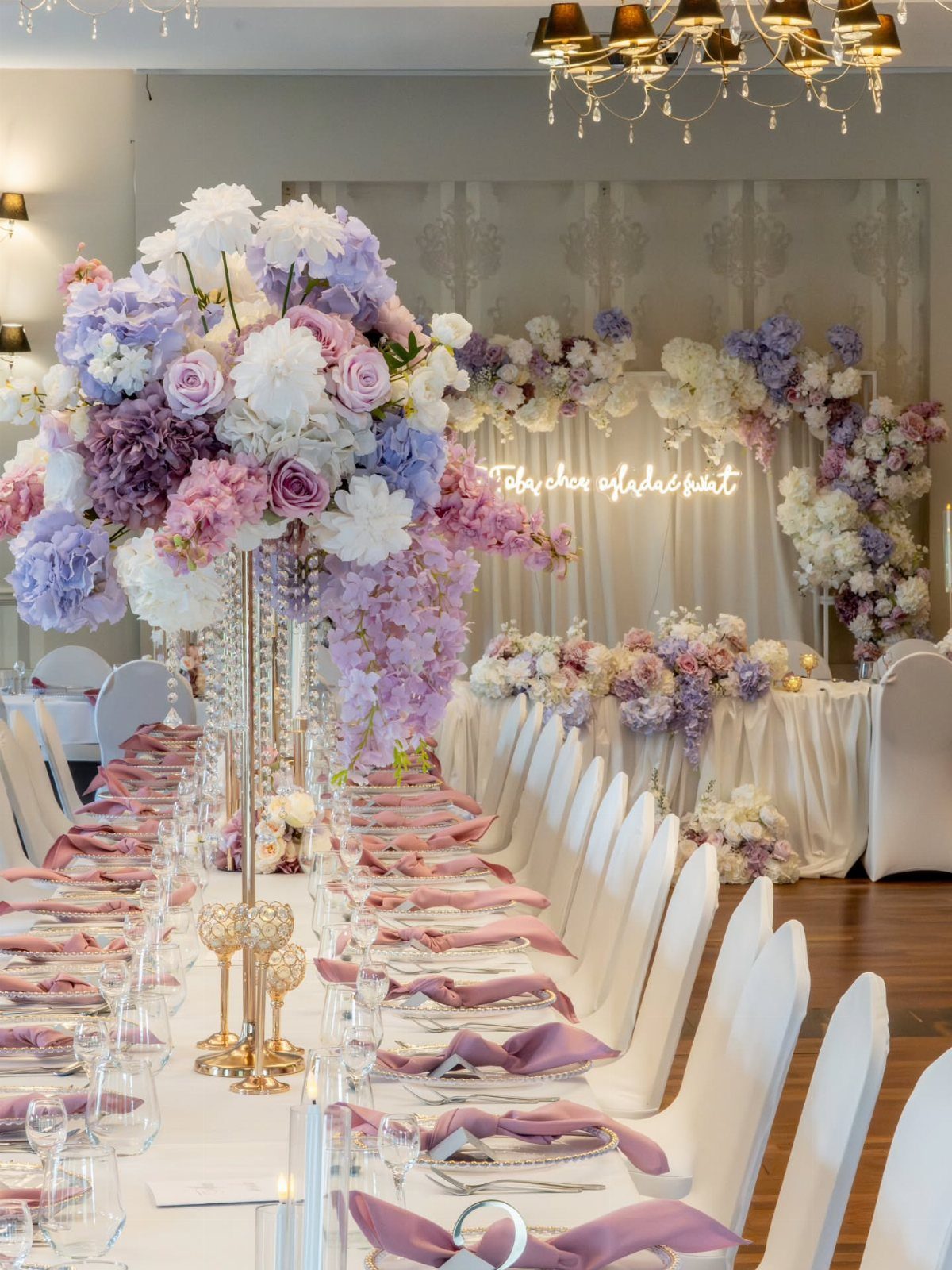 Tall lavender and white floral centerpieces on a long wedding reception table with pink napkins.