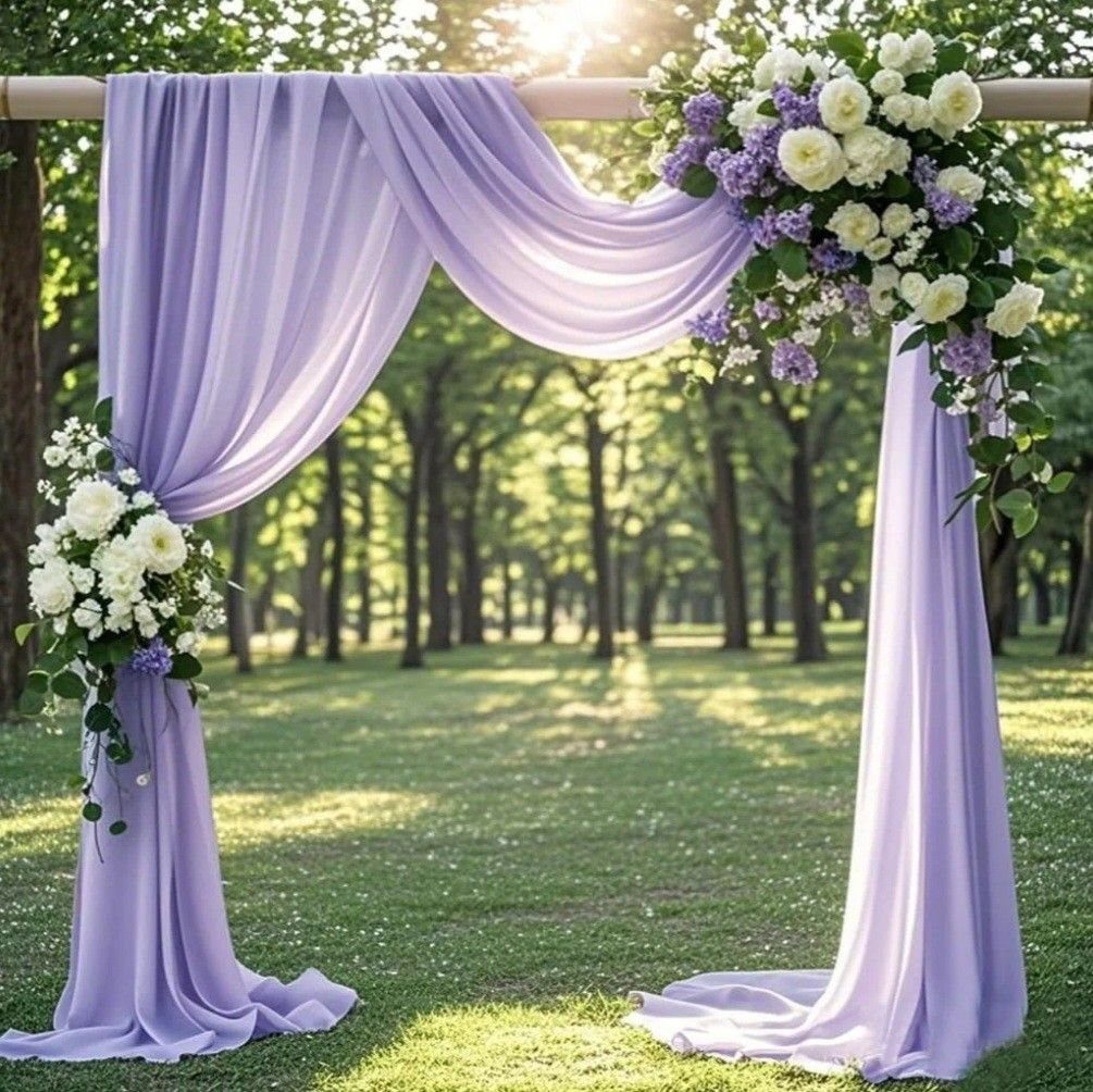 Lavender fabric draped over a wooden wedding arch decorated with white and purple flowers in a forest.
