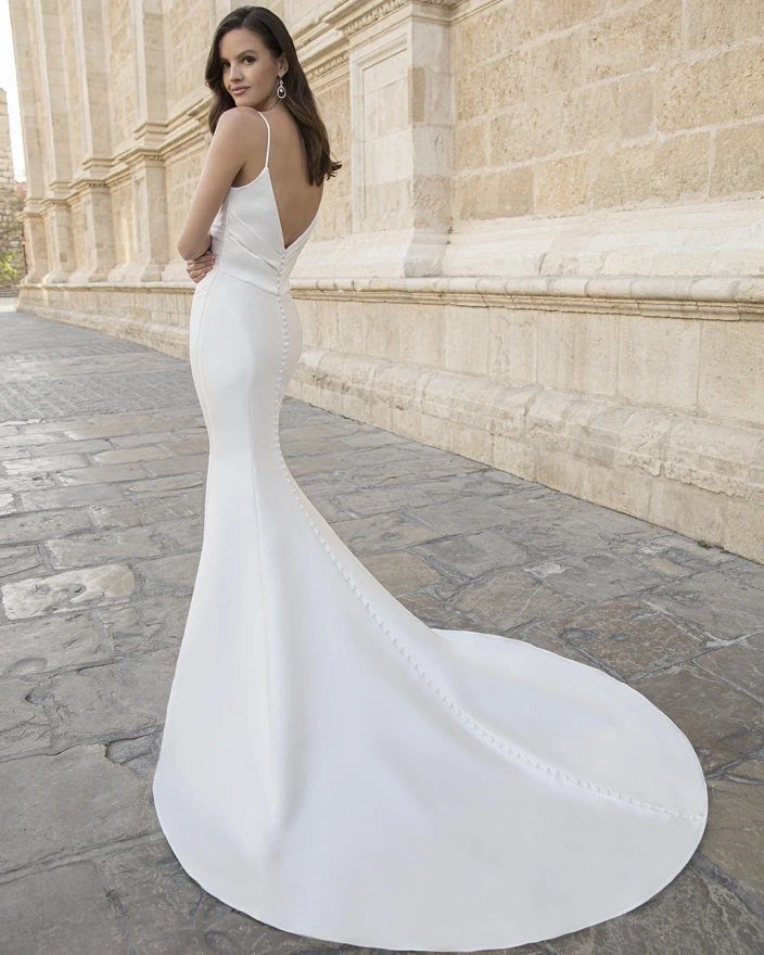 A woman wearing a sleek white satin wedding dress with a long train standing outdoors.
