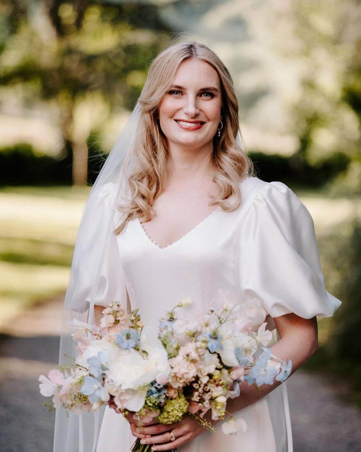 A smiling blonde bride wearing a white wedding dress with dramatic puffed sleeves holding a floral bouquet.