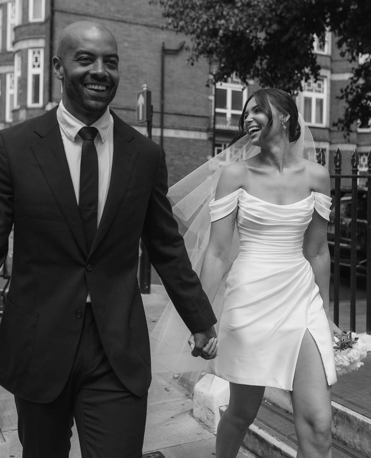 A bride in a short off-the-shoulder wedding dress walks hand-in-hand with her groom.