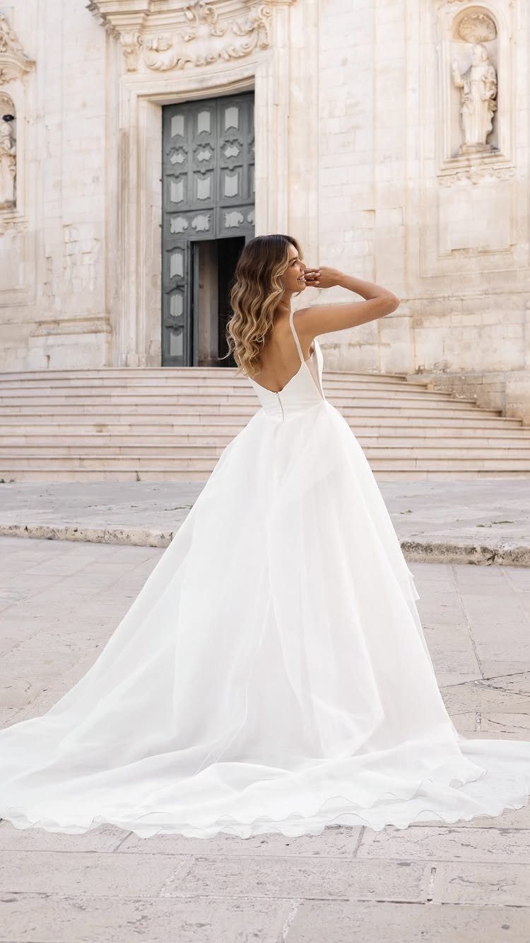 A woman wearing a white spaghetti strap wedding dress standing on stone steps outdoors.