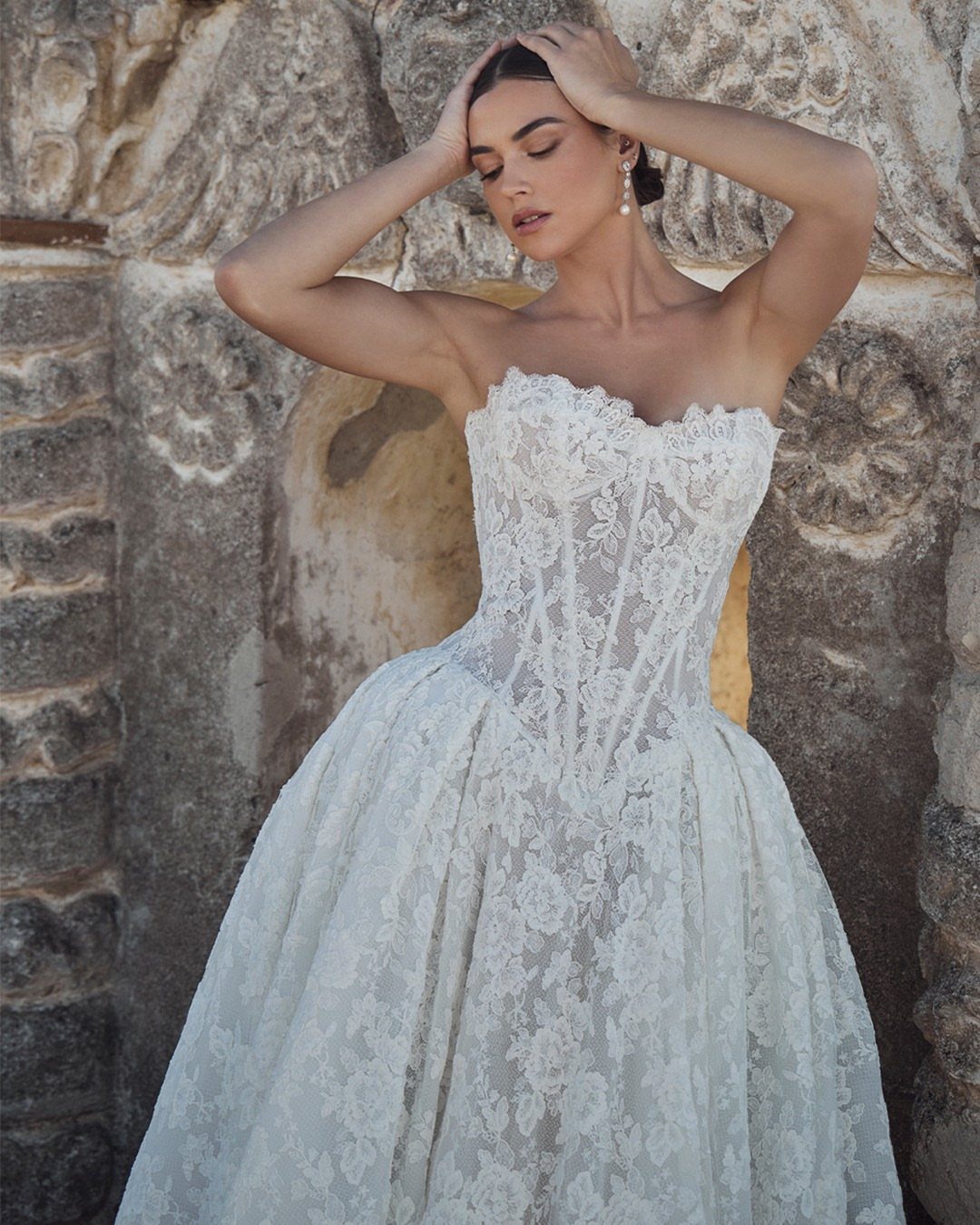 A woman wearing a white lace strapless wedding dress with a corset bodice against a stone wall.