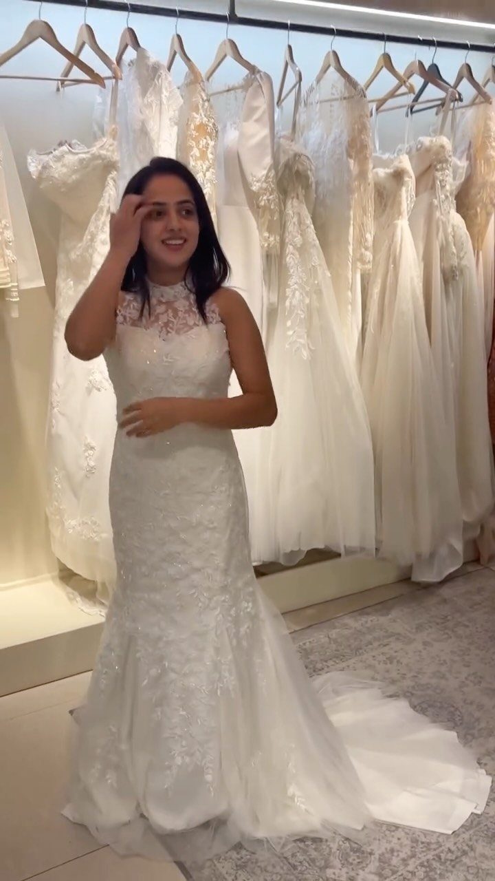 A smiling woman posing in a white lace mermaid wedding dress inside a bridal boutique.