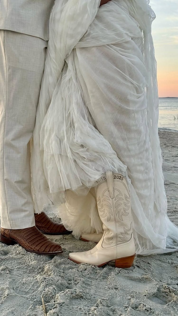 White embroidered cowboy boots worn under a flowing wedding dress on a sandy beach.