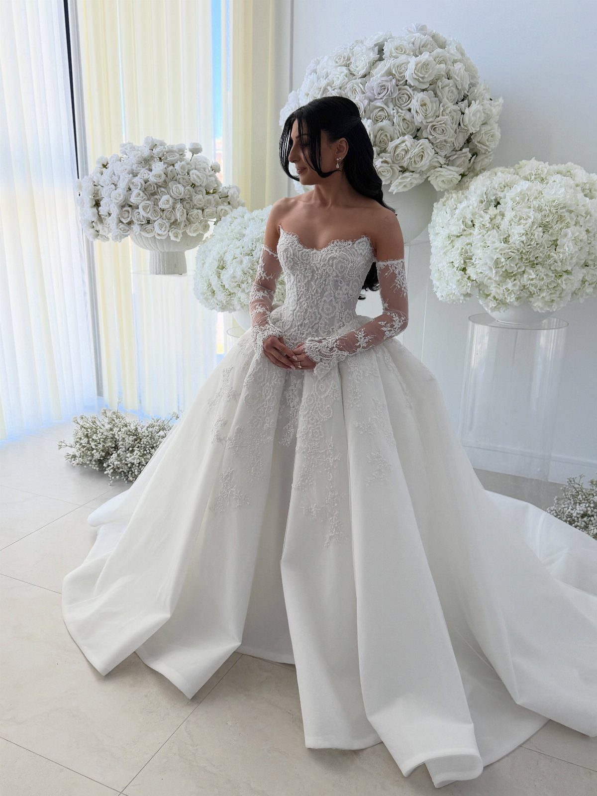 Woman in a white pearl-embroidered wedding dress posing among large white flower arrangements.