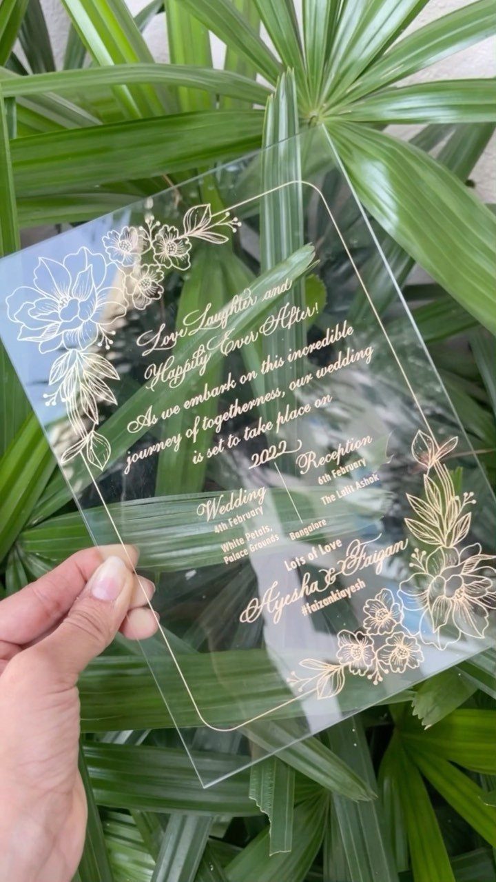 Wedding Invitations with Ribbon: 5 Ideas That Look Handmade 1 A hand holding a transparent acrylic wedding invitation with gold calligraphy and floral designs against green leaves.