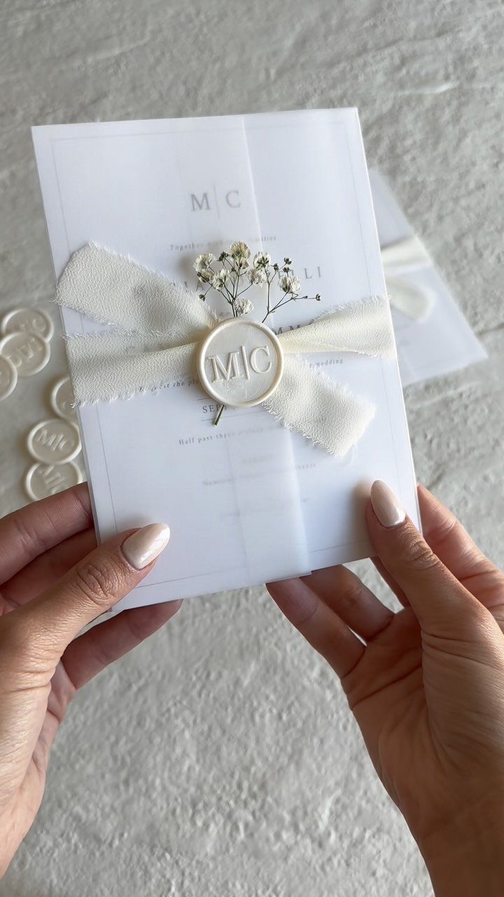 Wedding Invitations with Ribbon: 5 Ideas That Look Handmade 5 Hands holding a white wedding invitation decorated with a cream ribbon, wax seal, and dried flowers.