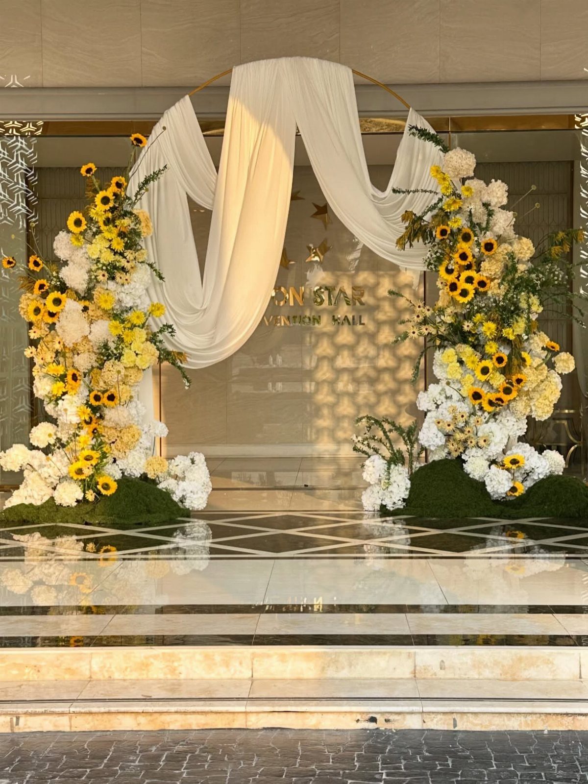 A circular gold wedding arch with white fabric drapes and large sunflower floral arrangements.