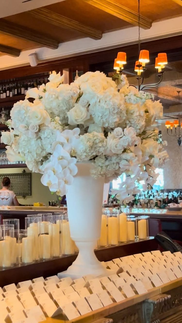 White Flower Centerpieces 5 Ways: From Lily Drama to Daisy Casual 1 Large white floral arrangement with roses and orchids in a white pedestal vase on a bar.