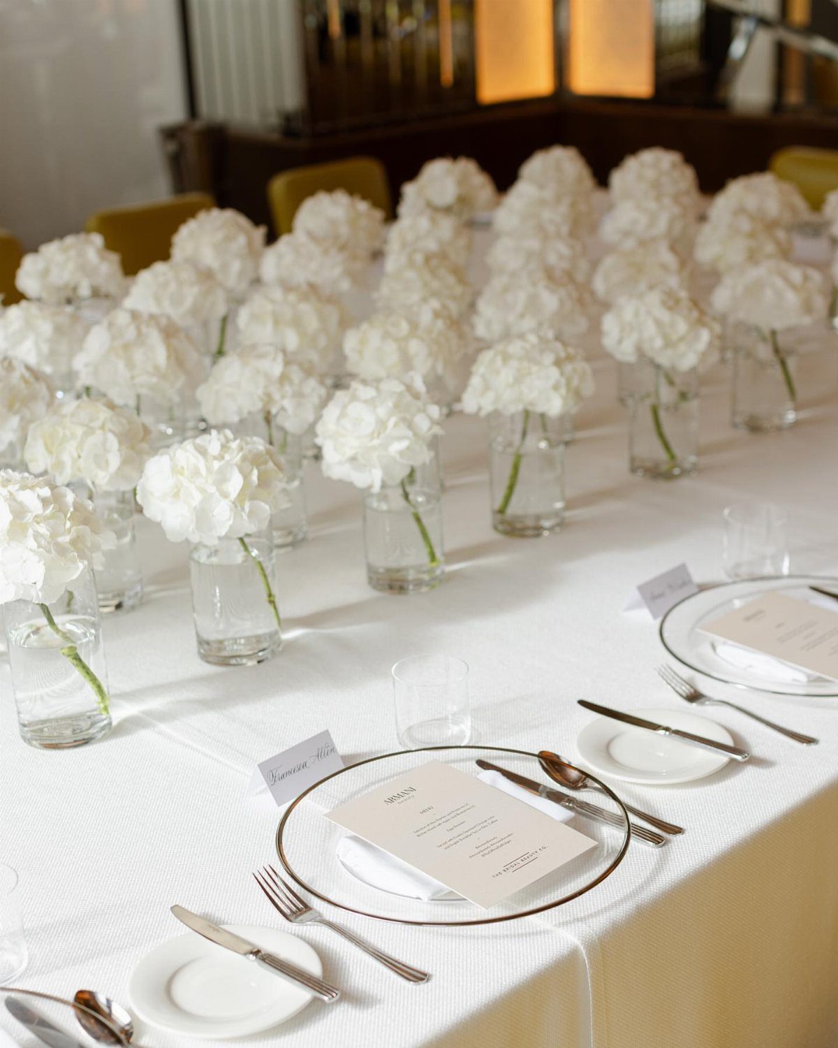 White Flower Centerpieces 5 Ways: From Lily Drama to Daisy Casual 4 Rows of small glass vases containing white hydrangea flowers on a long dining table.