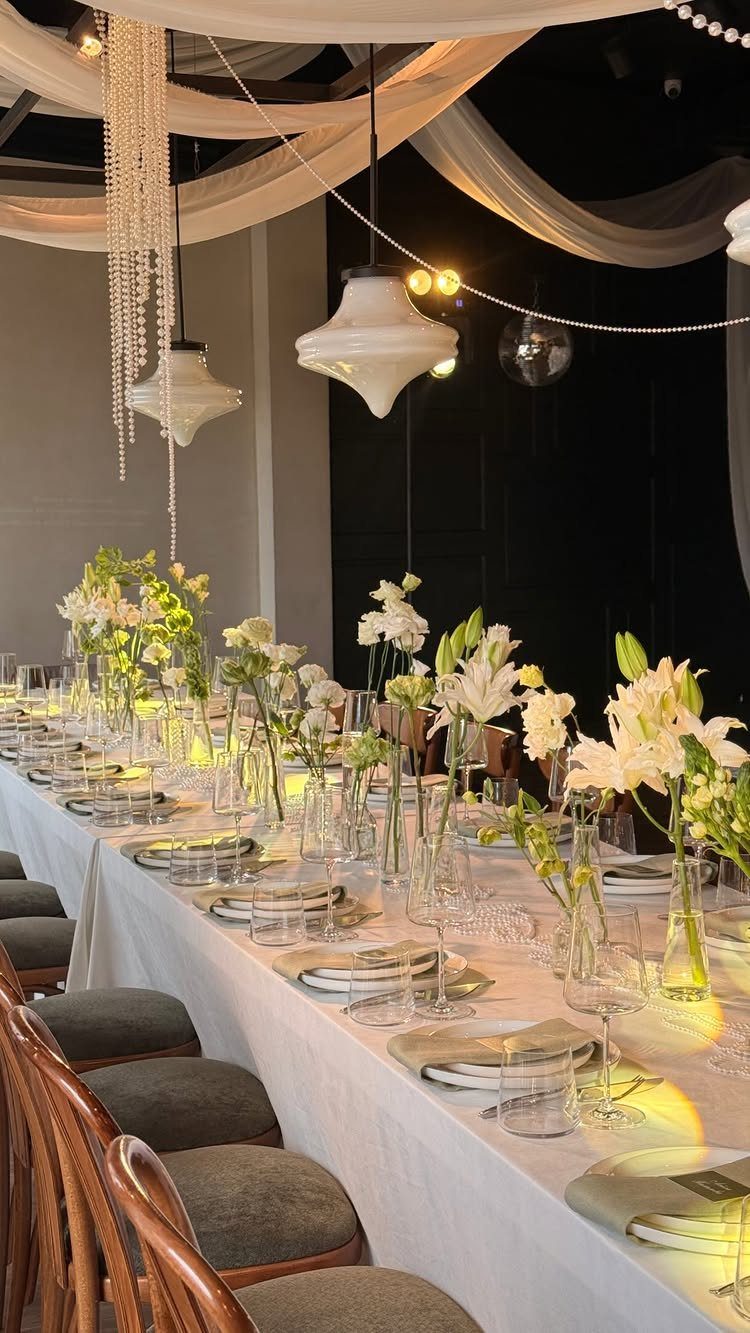 White Flower Centerpieces 5 Ways: From Lily Drama to Daisy Casual 2 White lily and rose centerpieces in glass vases lining a long, elegantly decorated banquet table.